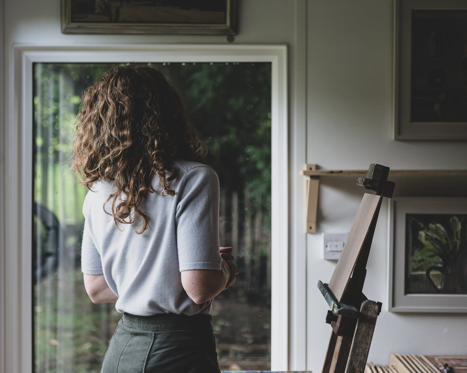 Sarah standing by a window looking outside, with a painting easel and framed picture in the foreground.