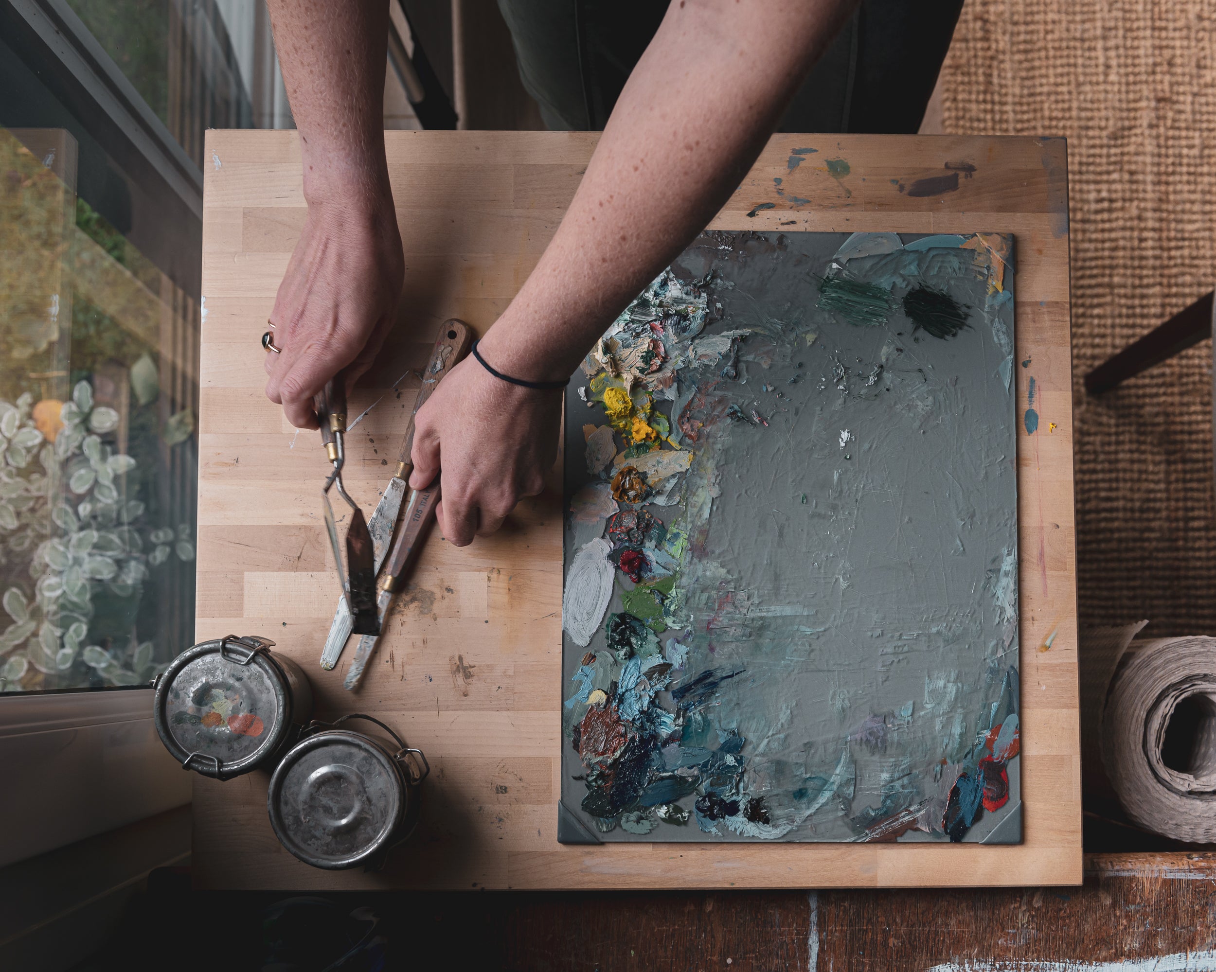 Sarah working on a painting with a palette and brushes on a wooden table.