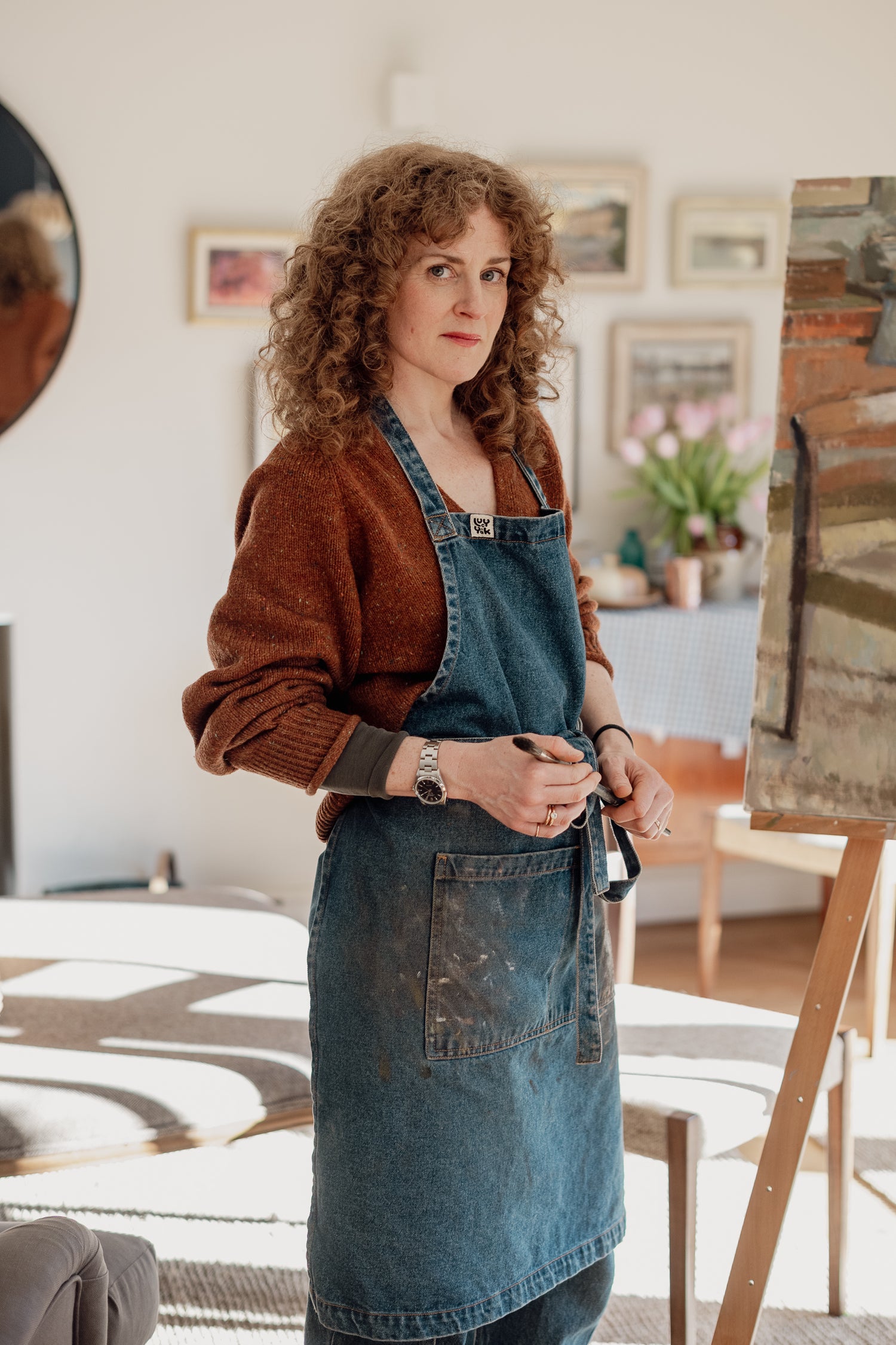 Sarah Manolescue in a denim apron standing in an artist's studio with art supplies and a rug.